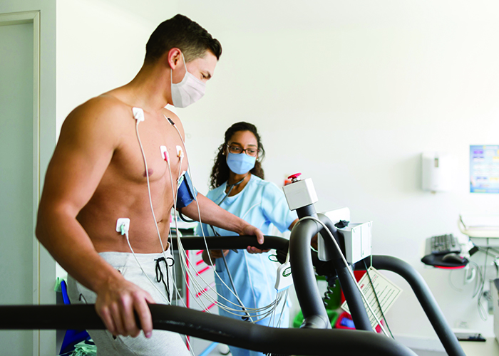 Masked male patient on a treadmill taking a stress test while a masked clinician looks on.