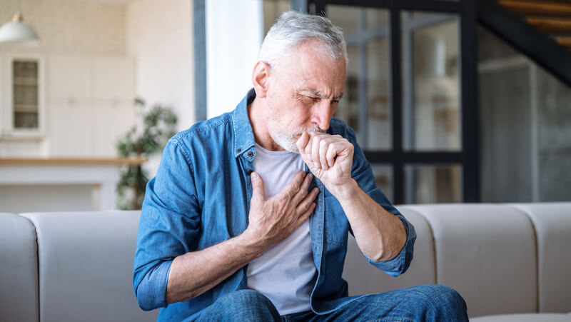 Grey-haired man coughs into his closed fist.